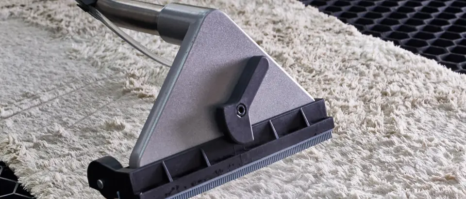A grey extraction wand with a steel tube and clear hose drawing water out of a wet cream shag rug on a black honeycomb mat