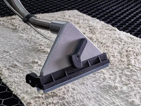 A grey extraction wand with a steel tube and clear hose drawing water out of a wet cream shag rug on a black honeycomb mat