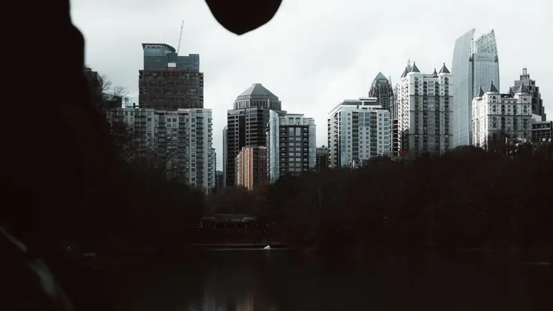 Midtown Atlanta towers seen across Lake Clara Meer in Piedmont Park under a grey sky, above dark trees and water and framed by leaves in silhouette
