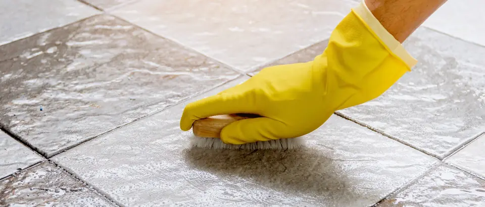 A hand in a yellow rubber glove scrubbing wet grey floor tiles with a wooden brush