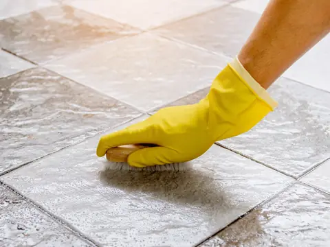 A hand in a yellow rubber glove scrubbing wet grey floor tiles with a wooden brush