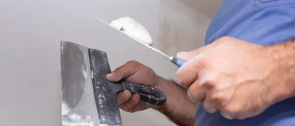 Two hands holding a wide taping knife and a smaller knife loaded with white filler in front of a smooth grey wall